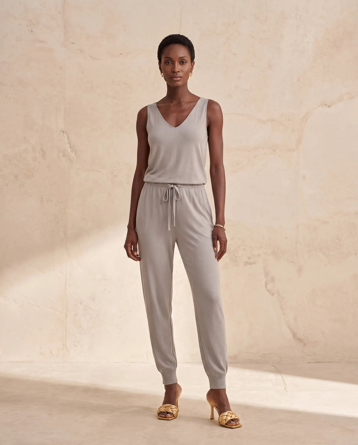 A woman stands against a warm beige textured wall wearing a light gray sleeveless V-neck jumpsuit with a drawstring waist, paired with gold heeled sandals.