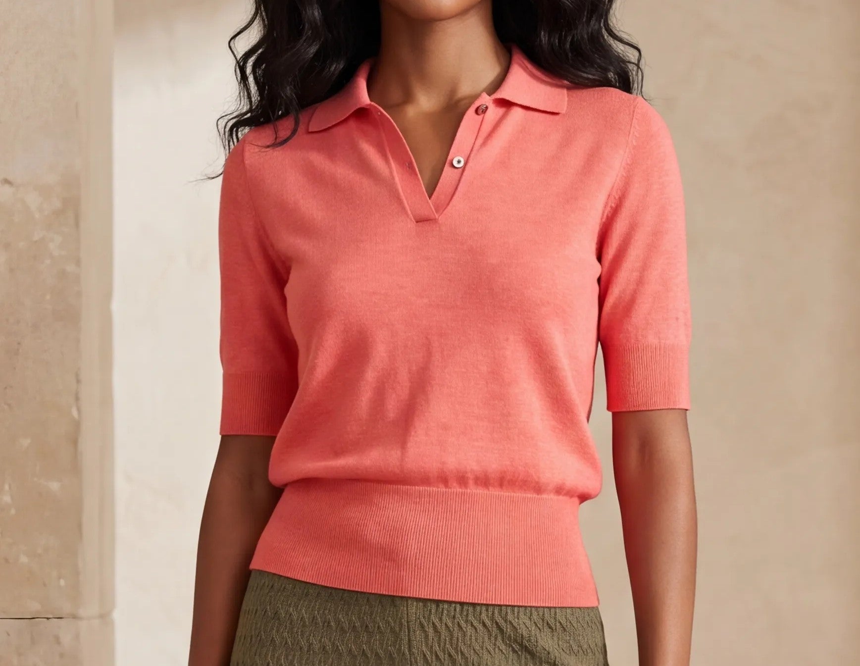 A woman wearing a coral pink short-sleeve knit polo shirt with a buttoned collar, paired with olive green textured pants, set against a warm beige background.