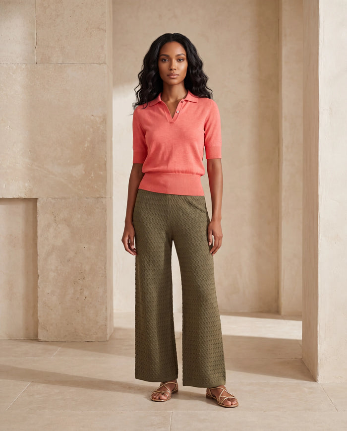 A woman stands in a warm, minimalist stone corridor wearing a coral pink short-sleeve knit polo shirt and textured olive green wide-leg trousers, paired with brown strappy sandals.