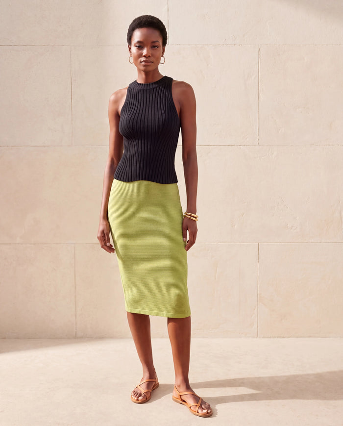 A model stands against a beige stone wall wearing a sleeveless black ribbed knit top and a lime green ribbed knit midi skirt, paired with tan strappy sandals.