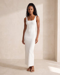 A woman modeling a knit sleeveless white maxi dress with a square neckline and thin shoulder straps, standing in a minimalist beige studio setting.